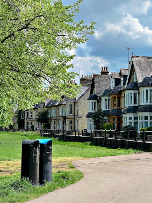 A row of traditional terraced houses with bay windows and slate roofs, situated along a residential street. In the foreground, a grassy area is bordered by a paved footpath and a black metal fence. Tw
