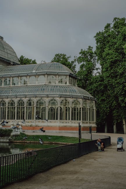An expansive, ornate greenhouse structure with a tall, curved glass ceiling supported by white metal framework and decorative columns. The glass panels form a series of arched windows and intricate pa