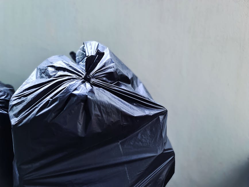 A close-up view of a tightly knotted black plastic rubbish bag resting against a plain, light-colored wall. The bag appears to be made of glossy, durable plastic material with visible creases and ripp