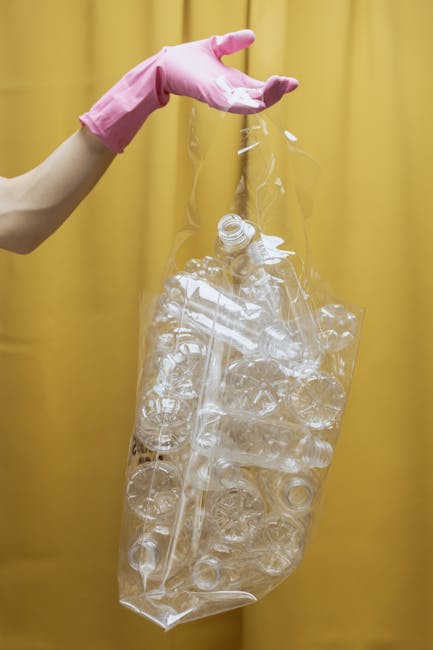 A close-up view of a tightly knotted black plastic rubbish bag resting against a plain, light-colored wall. The bag appears to be made of glossy, durable plastic material with visible creases and ripp