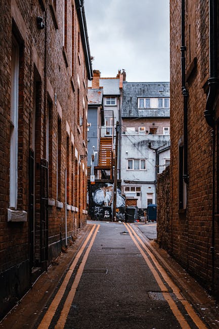 A narrow alleyway between two brick buildings in an urban setting, with a paved surface marked by double yellow lines along each edge. The left building features red brick walls with small, evenly spa