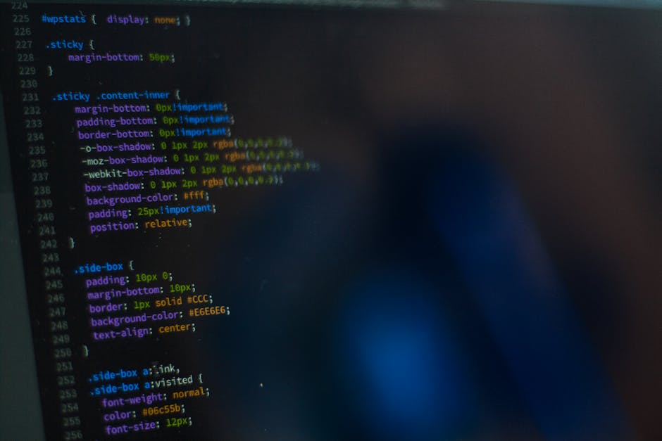 Close-up view of a computer screen displaying lines of HTML code in a dark-themed editor, with syntax highlighting in various colors such as green, orange, and white. The code includes HTML tags like 